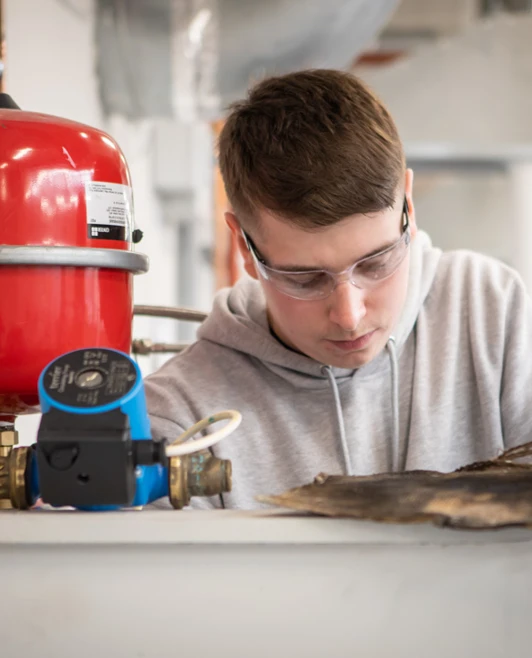 A plumbing student wearing safety glasses, closely inspecting and working on a heating system setup with copper pipes and a red expansion vessel. A plumbing student wearing safety glasses, closely inspecting and working on a heating system setup with copper pipes and a red expansion vessel.